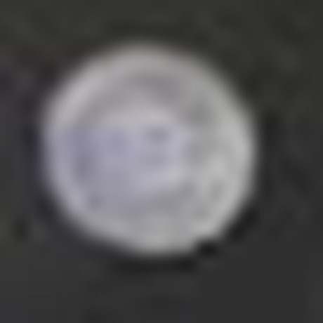 A heavily enlarged, blurred crop of a small round pale badge on a dark jacket lapel; no design is discernible.
