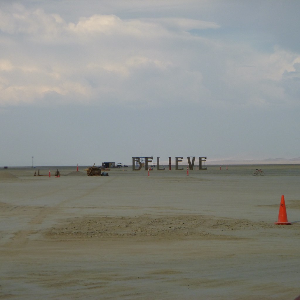 Large letters spelling BELIEVE stand on an open desert playa beneath a cloudy sky.