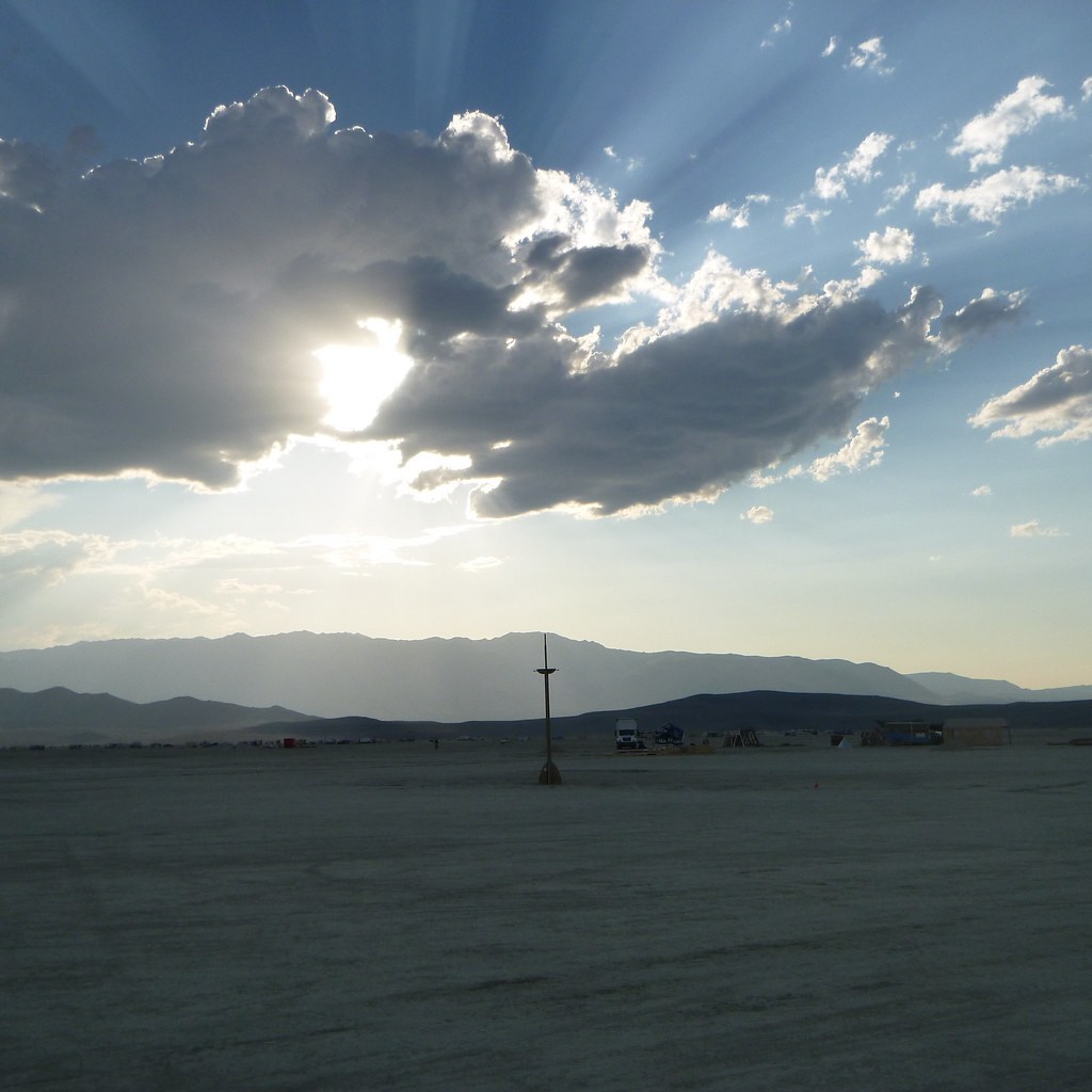 Sunlight streams around a dark cloud over a flat desert landscape and distant mountains.