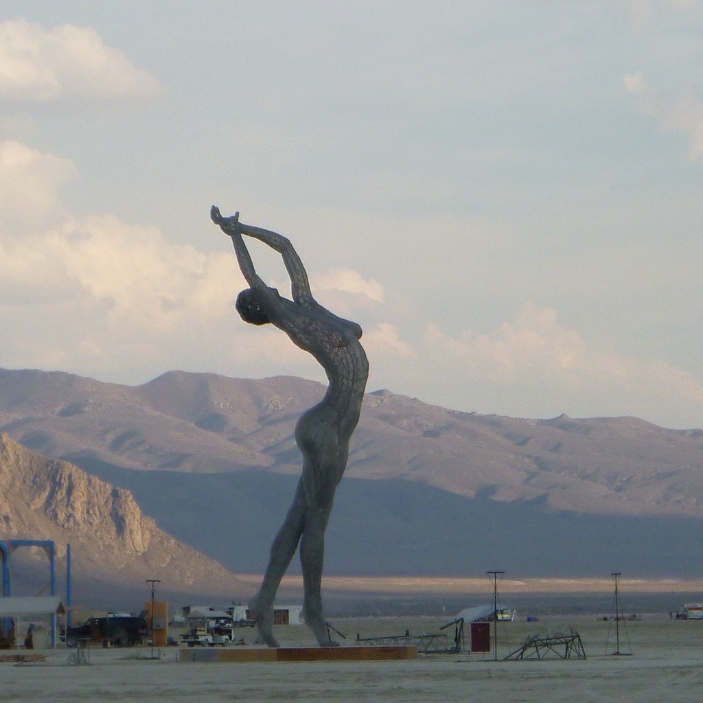 A tall, curved human-form sculpture stands on the desert floor with mountains behind it.