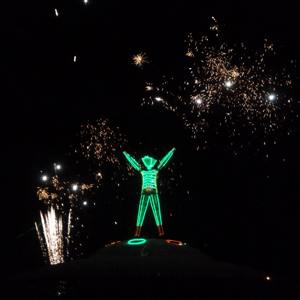 A green illuminated figure on a circular platform beneath bright fireworks.