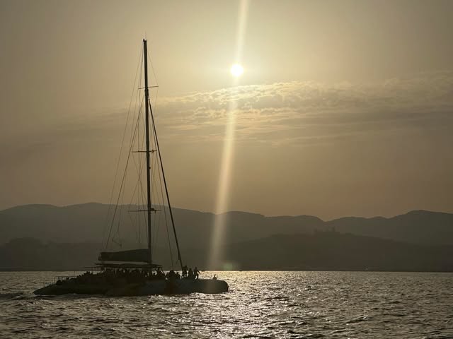 A sailboat rests on sunlit water with dark hills and a low golden sun beyond.