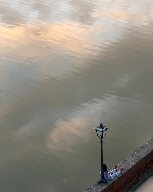 Soft clouds reflected in river water beside a streetlamp and stone embankment.