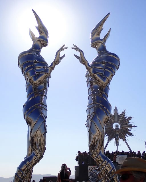 Two tall sculptural figures in patterned armor reach toward each other beneath pointed headdresses.